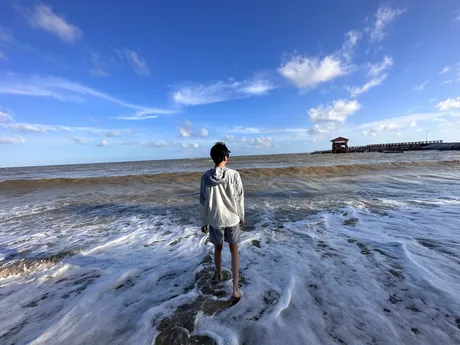 A figure standing in shallow surf under a wide blue sky